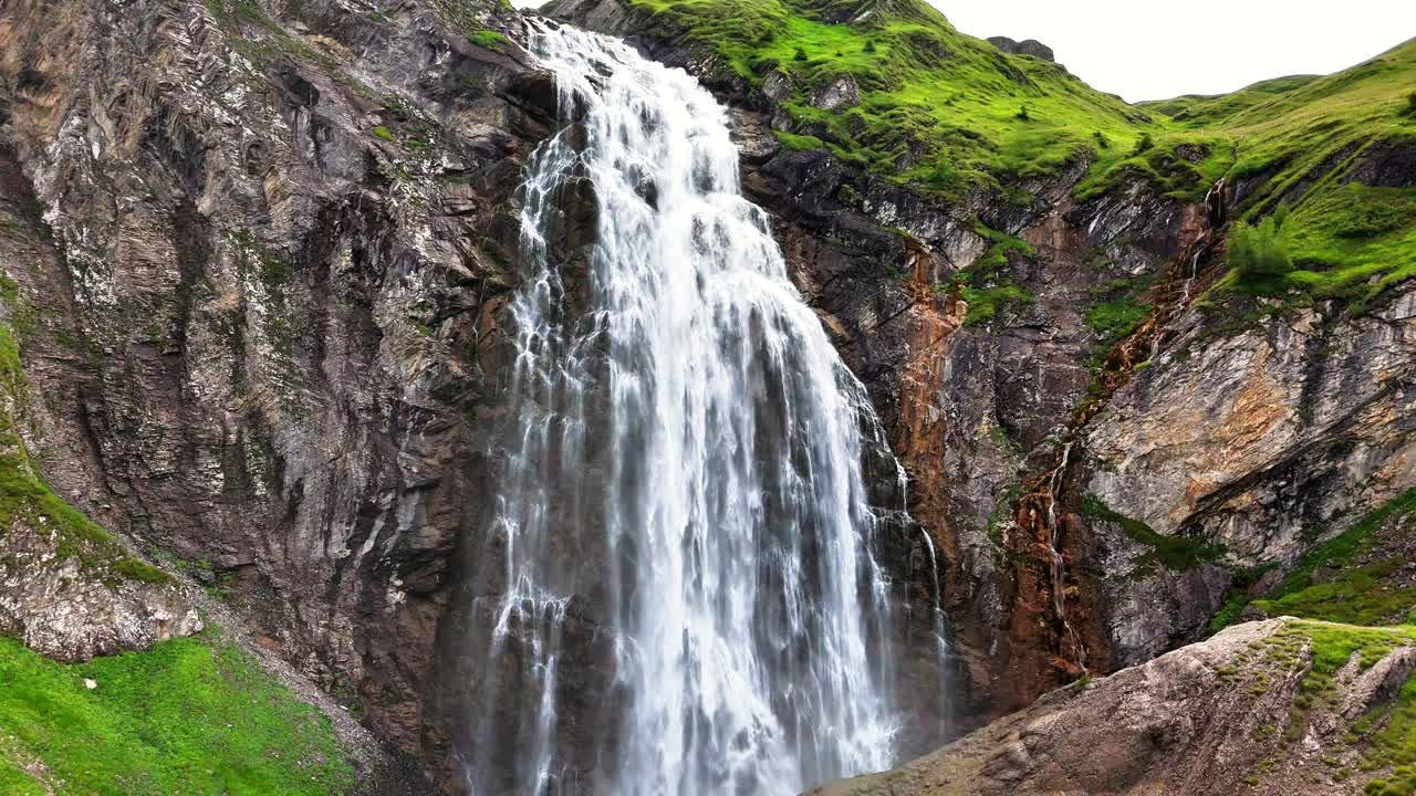 Engstligen waterfalls cascading down a rugged mountain, flowing over rocks, green grass, creating a powerful and scenic natural spectacle in Adelboden, Switzerland, drone pulling away