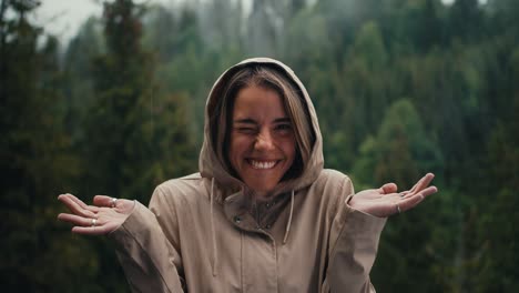 chica rubia feliz se regocija en la lluvia del bosque en las montañas divirtiéndose y mirando a la cámara
