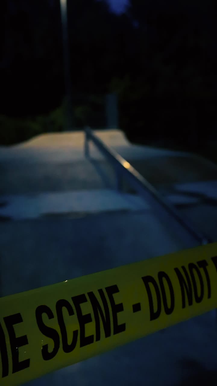 Crime scene yellow colored tape ribbon pulled in front of a concrete skatepark where metallic rail is visible that is reflecting street lights during dark evening with bokeh blurry background