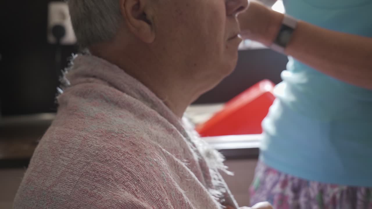 Head profile of grey haired old man having haircut in home kitchen by amateur barber, left to right slide close up.