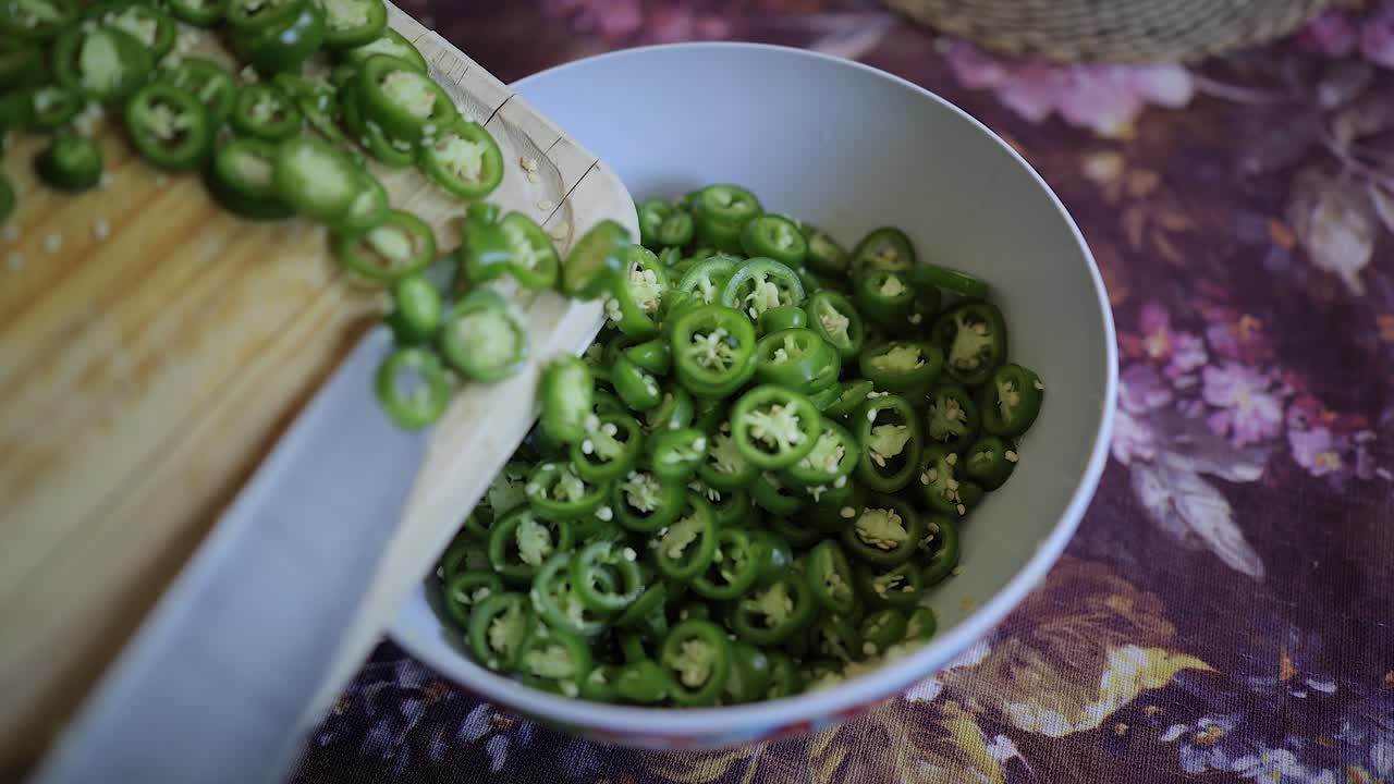 Sliced jalapeno peppers from cutting board slide into kitchen bowl
