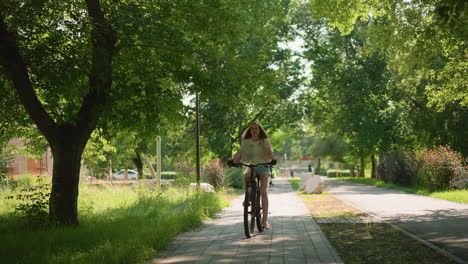 lady confidently riding bicycle along a lush, green park path, her long hair flowing freely, the background includes tall trees, street lamps, a building, and people walking in the distance