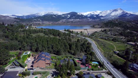 Smooth-cinematic-push-in-aerial-over-the-town-of-Navacerrada-towards-the-reservoir-and-the-snow-capped-mountains