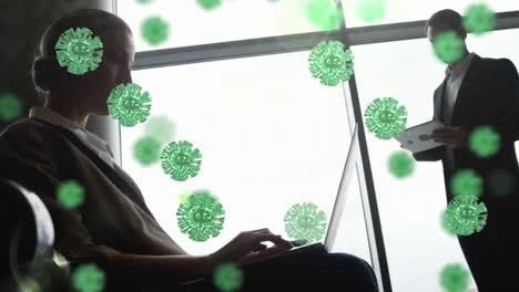 Animation-of-floating-Covid-19-cells-over-Caucasian-man-and-woman-using-tablet-and-laptop-at-airport