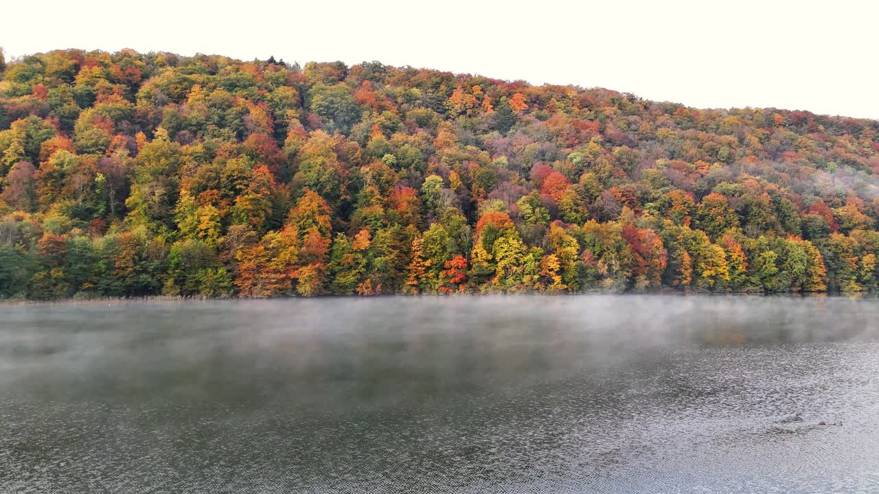Mist drifts above Lac du Val as autumn colors blanket the forested hillside near Le Herisson in France. Vibrant foliage in orange, red, and green surrounds the calm lake, creating a seasonal landscape