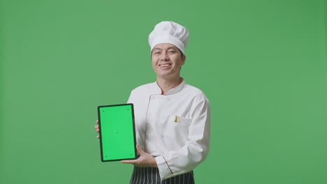el chef asiático sonriendo y mostrando la tableta de pantalla verde a la cámara mientras está de pie en el estudio de fondo de la pantalla verde