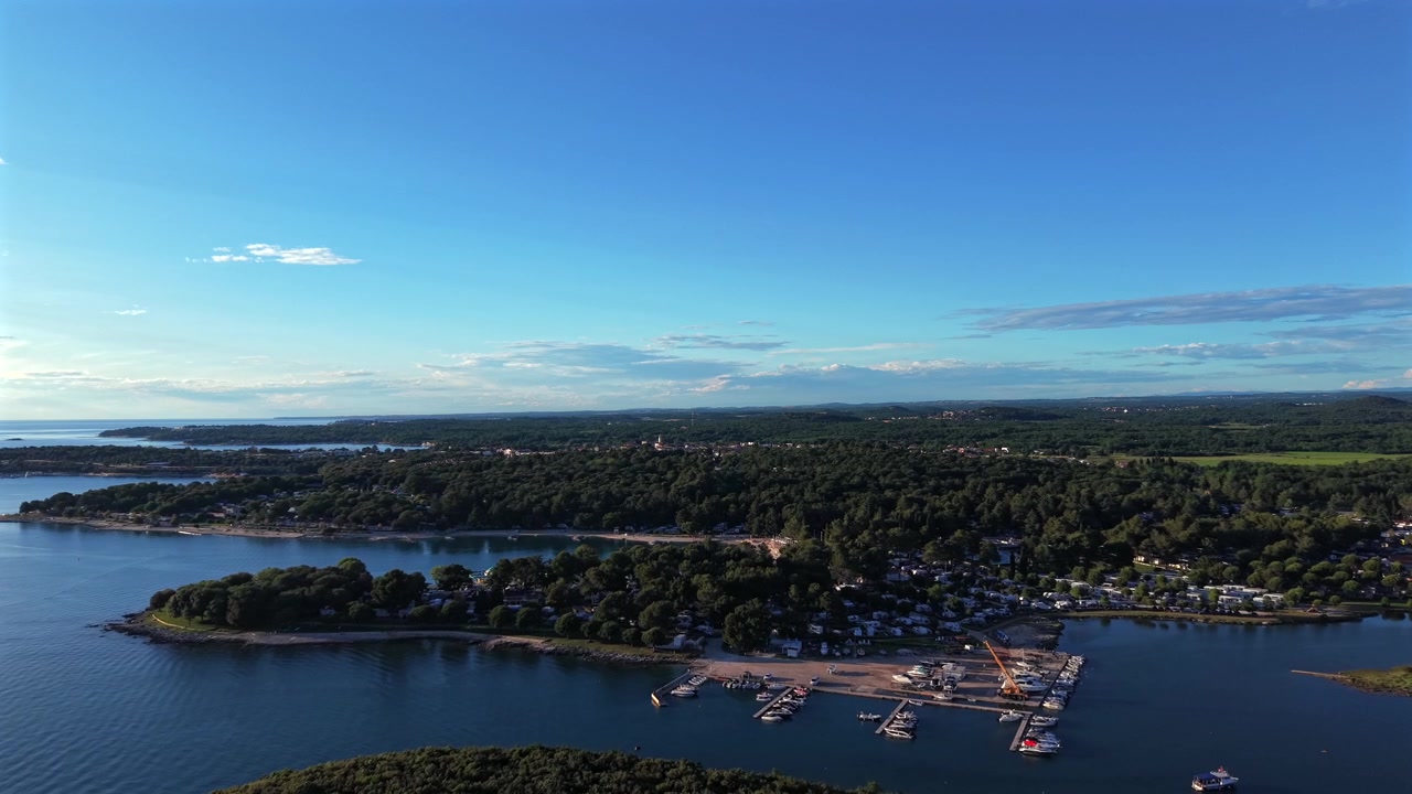 Forested coastal marina hosts docked boats near lakeside camping site in Istria, Croatia, calm blue bay winds past tree-covered islands beneath golden evening summer sky. Aerial drone approaching shot