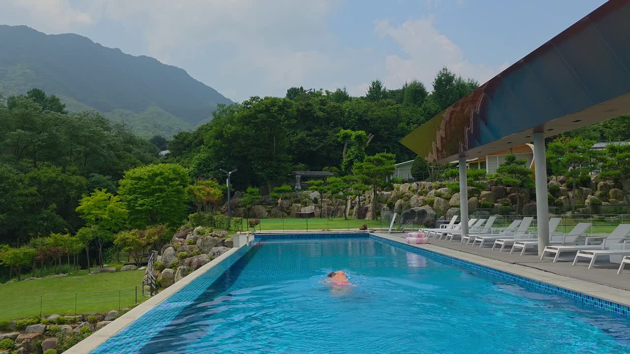 A man in red swim trunks dives into the water and swims along the scenic infinity pool at Glamtree Resort, surrounded by lush greenery and mountain views in Gapyeong, South Korea