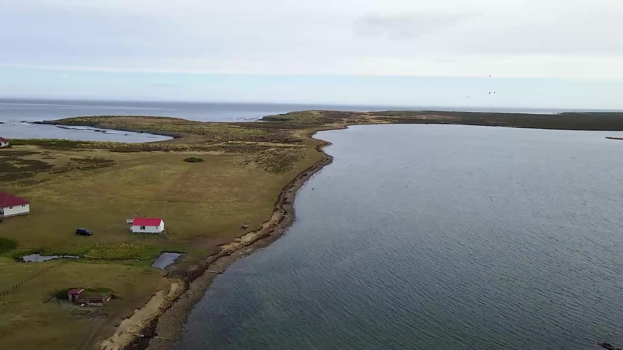 La telecamera panoramica su un'isola remota con pochi edifici, rivelando la vastità dell'oceano circostante.