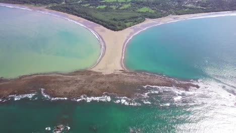 aerial shot pulling away from the whale tail shaped rocky point of punta uvita surrounded by bright blue ocean waves in south puntarenas, costa rica