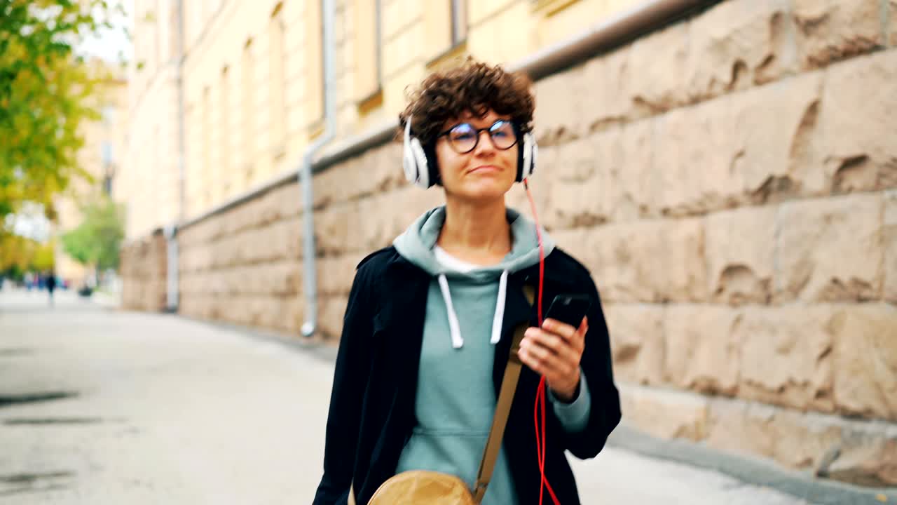 Cheerful young lady is listening to music through headphones and holding smartphone walking in street alone on autumn day. Youth culture and technology concept.