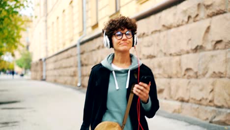 cheerful young lady is listening to music through headphones and holding smartphone walking in street alone on autumn day. youth culture and technology concept.