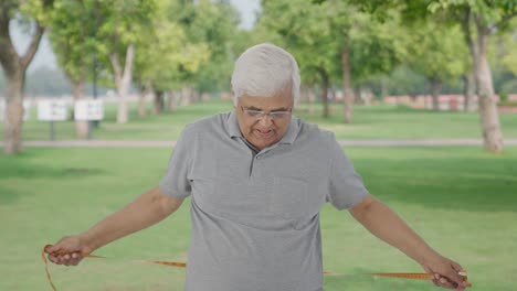 Sad-Indian-old-man-checking-his-waist-size-using-inch-tape-in-park