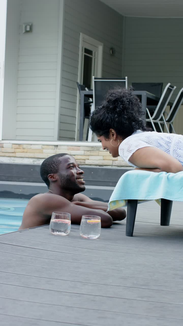 Vertical video: Couple talking, woman coming, leaning poolside, man smiling with drinks, copy space