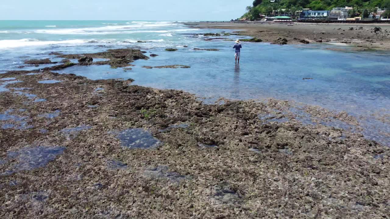 volando sobre las piscinas de marea de la ciudad junto a la playa con un tipo mirando hacia la playa de pipa del océano, brasil por un dron 4k