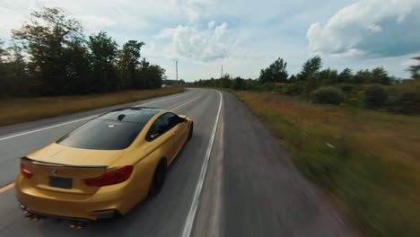 fpv drone video following gold bmw m4 sports car along winding country road through woods