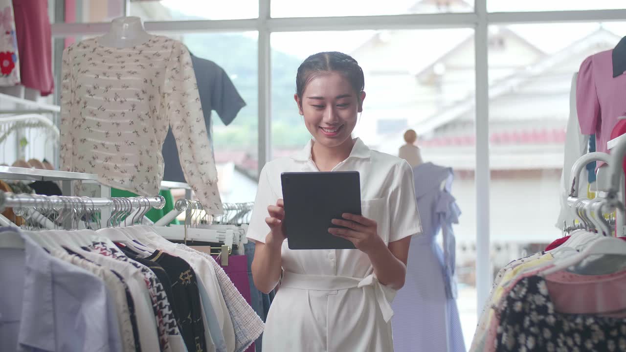 태블릿을 사용하는 옷 가게의 아시아 여성 주인이 레일에 있는 재고를 확인하는 동안 카메라에 미소 짓고 있습니다.