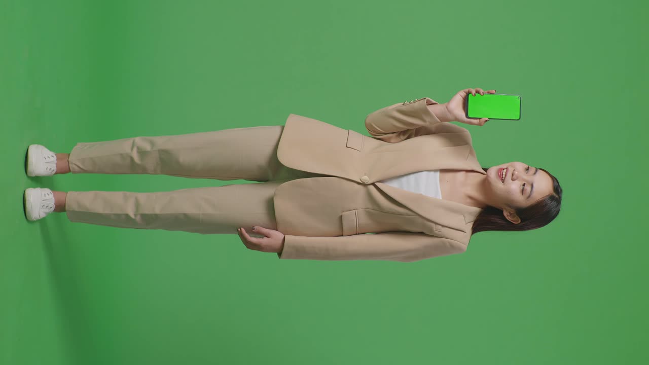 cuerpo lleno de mujer de negocios asiática con una sonrisa que muestra pantalla verde teléfono móvil mientras está de pie frente a la pantalla verde