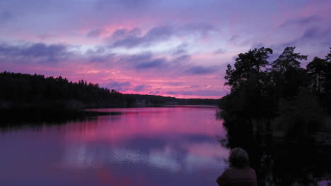 aerial drone view bypassing a man sitting at a rocky shore, of a lake, a purple sky, at a colorful sunset or dusk, at albysjon, tyreso, sweden