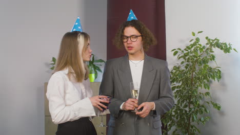 Niño-Y-Niña-Con-Sombrero-De-Fiesta-Tomando-Bebidas-Y-Hablando-Juntos-En-La-Fiesta-De-La-Oficina