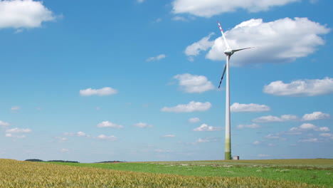 low drone shot over farm field of tall wind turbine against blue cloudy sky