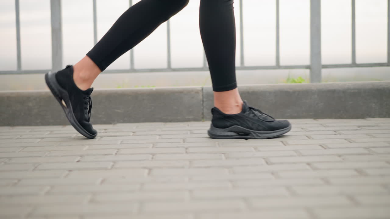 Close-up leg view of girl walking confidently in black canvas shoes on interlocked pavement, with iron fence beside her, showcasing casual outdoor activity with a peaceful urban backdrop