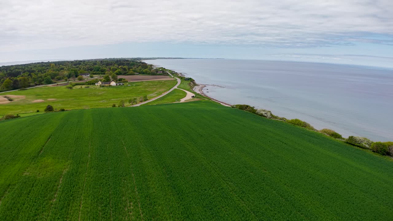 Aerial drone footage of Denmark’s coastal farmland with green fields, gentle hills, and shoreline meeting calm blue water under bright daylight