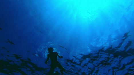 Einzigartige-Unterwasserperspektive-Eines-Fitten-Mannes,-Der-In-Den-Blautönen-Des-Ozeans-Schwimmt,-Während-Sonnenstrahlen-Durch-Das-Wasser-Gehen
