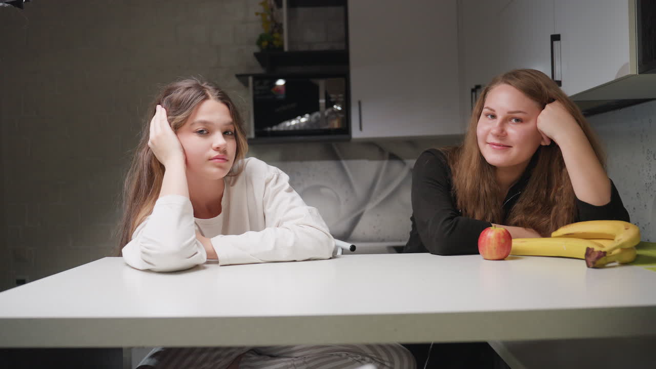 Buddies seated at kitchen table resting chin on hands admire each other with warm expressions while occasionally looking away, apple and bananas on surface, relaxed indoor atmosphere