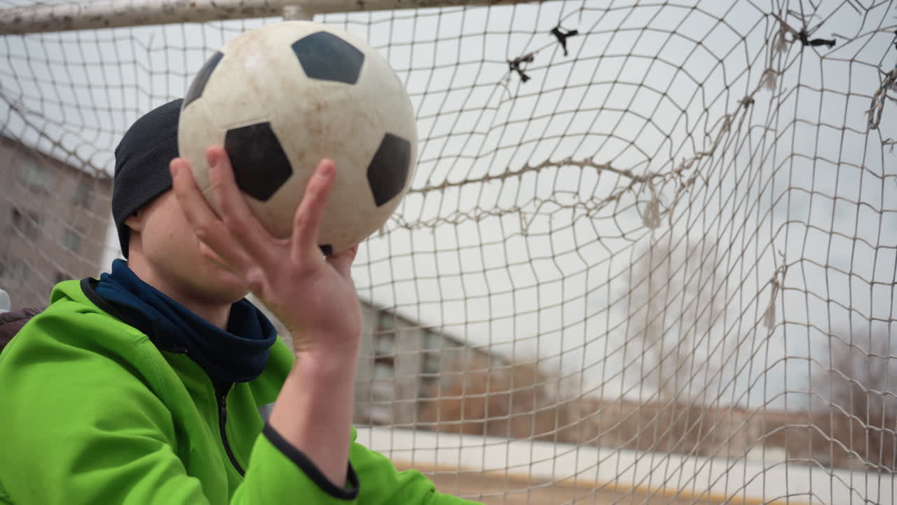 흐린 날씨 속에서 집중한 표정으로 공을 멋지게 잡아내는 도시 골키퍼. 초록색 코트와 비니를 착용한 노련한 백인 남성이 흐린 날 축구 경기 중 공을 성공적으로 잡아냅니다.