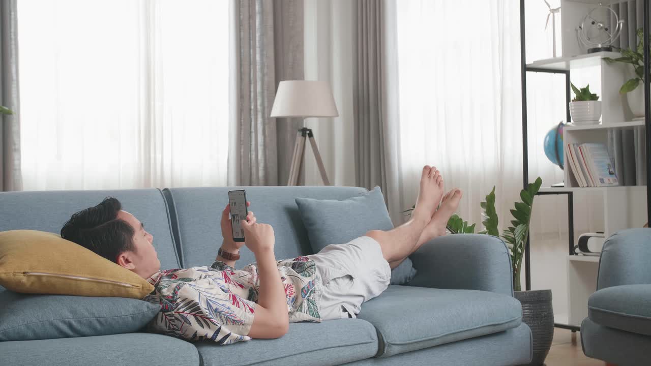 Asian Man Lying On Couch In Living Room And Using Smartphone Buy Fashion Clothes Online, Online Shopping And Lifestyle Concept