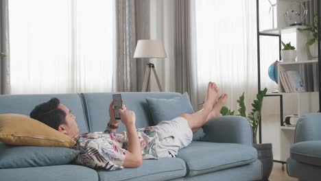 asian man lying on couch in living room and using smartphone buy fashion clothes online, online shopping and lifestyle concept