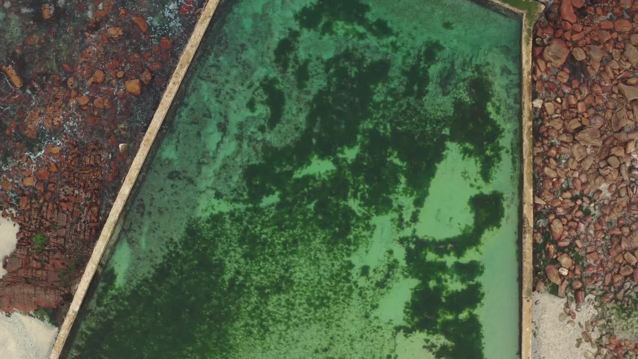 Top-down drone shot captures a green tidal pool along the shore at a beach in Greece. Contrast between the pool and sandy shore creates a striking view.