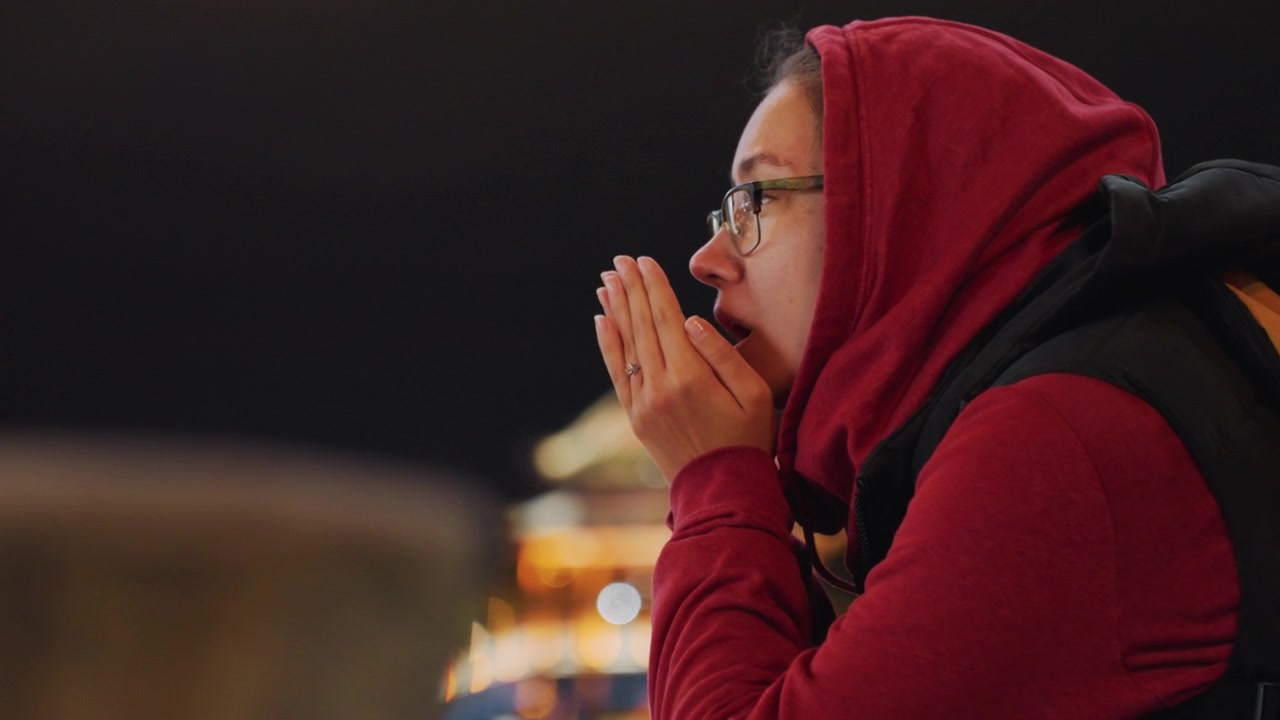 Pemandangan Tepi Laut Malam Hari, Pengamat Wanita dengan Tangan Terkatup Penuh Harap di Dekat Wajah Menatap Arsitektur yang Diterangi, Pantulan Lembut di Air, Suasana Meditatif yang Tenang, Syal dan Ransel Mengisyaratkan Perjalanan