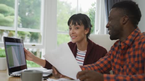 Happy-diverse-couple-sitting-at-table-talking-and-working-with-laptop