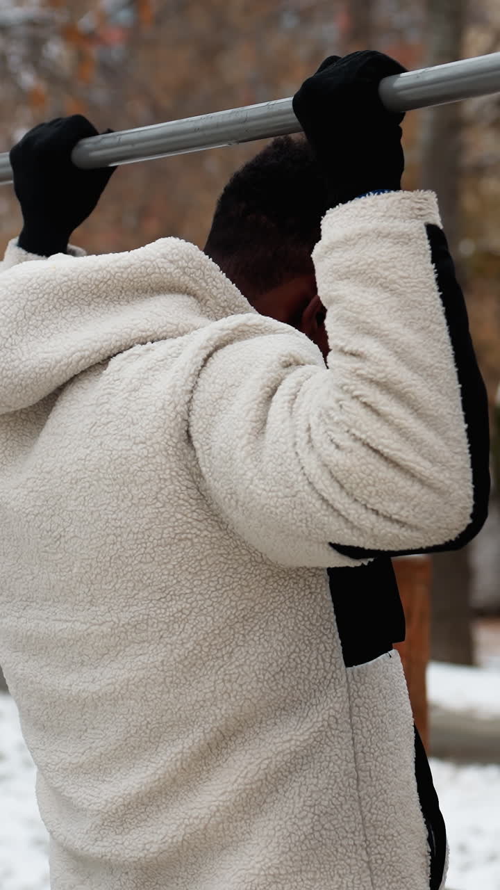 겨울 재을 입은 젊은 성인이 눈 인 공원 배경으로 야외 운동 장비의 철 바를 잡기 위해 점프하여 운동을 위해 따뜻해지는 모습