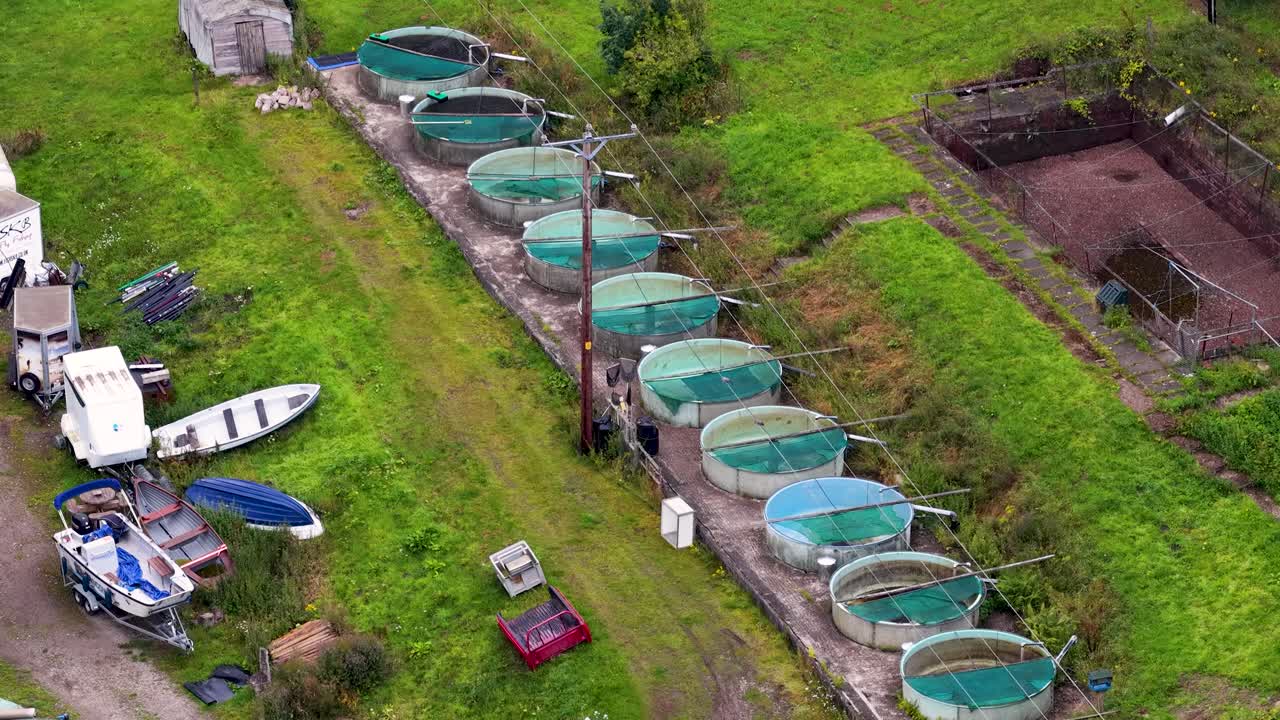 Aerial footage moves over circular aquaculture tanks, boats, and equipment in a green rural setting, under natural daylight, with smooth camera motion