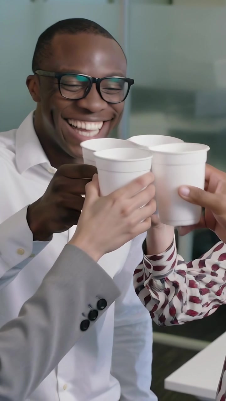 Vídeo vertical: Homem rindo, de óculos e camisa branca, liderando a equipe batendo copos para um brinde no escritório