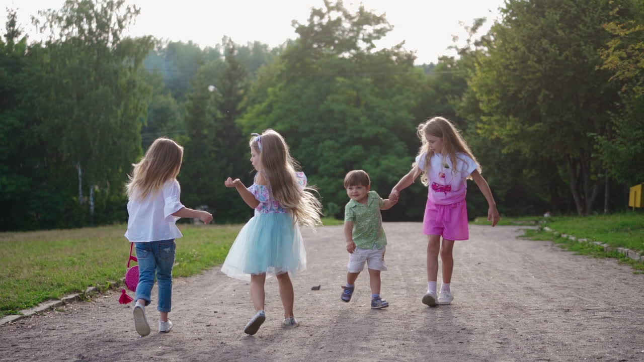 Joyful children on the background of green trees in summer. Girls and little boy walking merrily in the park. Happy childhood.