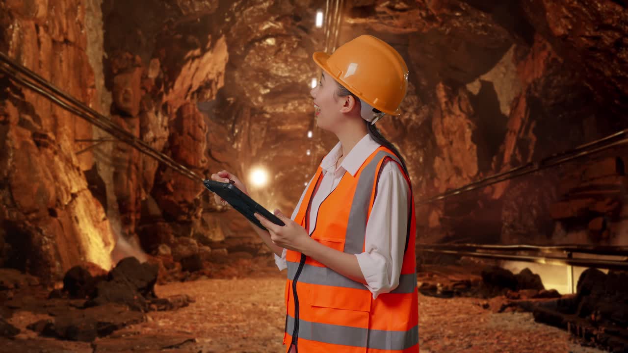 Side View Of Asian Female Engineer With Safety Helmet Taking Note On The Tablet And Looking Around In Underground Mine Tunnel