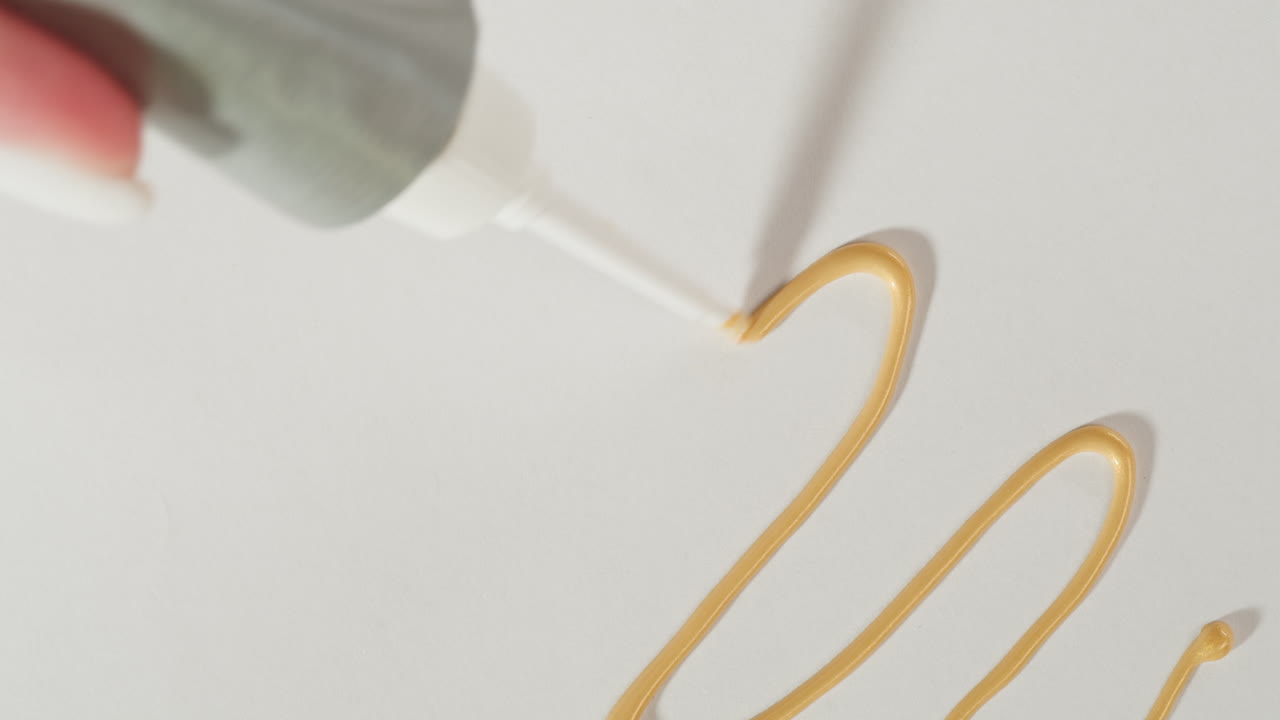 Close-up of Hand Squeezing Golden Liquid from Tube onto White Surface