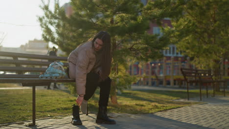 a girl in a peach jacket and black trousers is sitting on a bench in a park. she is seen taking off one boot while the rollerblades rest on the bench beside her