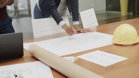 Diverse-architects-inspecting-blueprints-on-table-with-helmet-in-office,-in-slow-motion