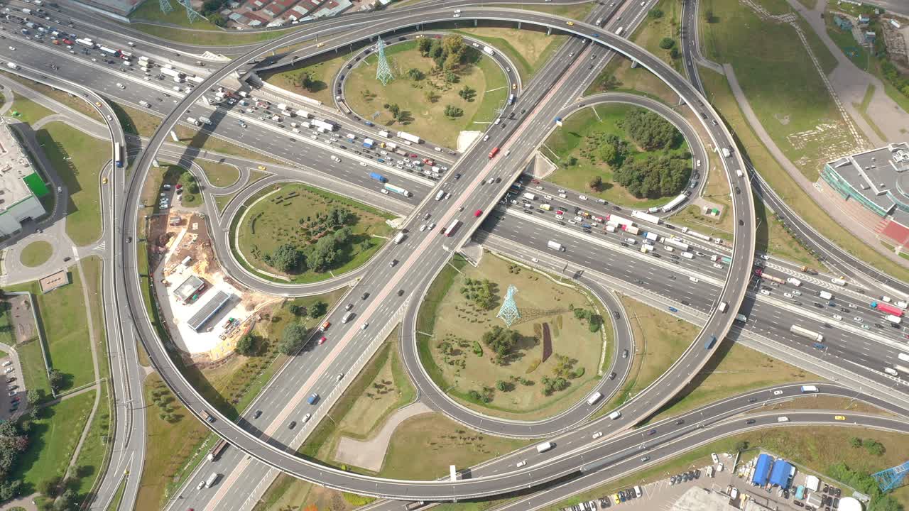 vuelo alrededor de la gran intersección circular con vueltas, curvas y salidas de trébol. vista aérea de arriba hacia abajo desde el avión no tripulado. luz solar y sombra de las nubes. áreas verdes y edificios alrededor. la cámara se mueve alrededor del punto de vista y mirando el tráfico..