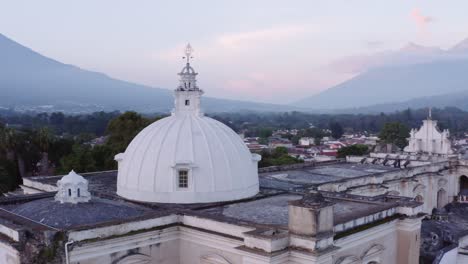 Dynamic-drone-shot-orbits-horizontally-around-the-white,-round-roof-of-San-Francisco-the-Great-Sanctuary-in-Guatemala,-revealing-Agua-volcano,-near-Antigua