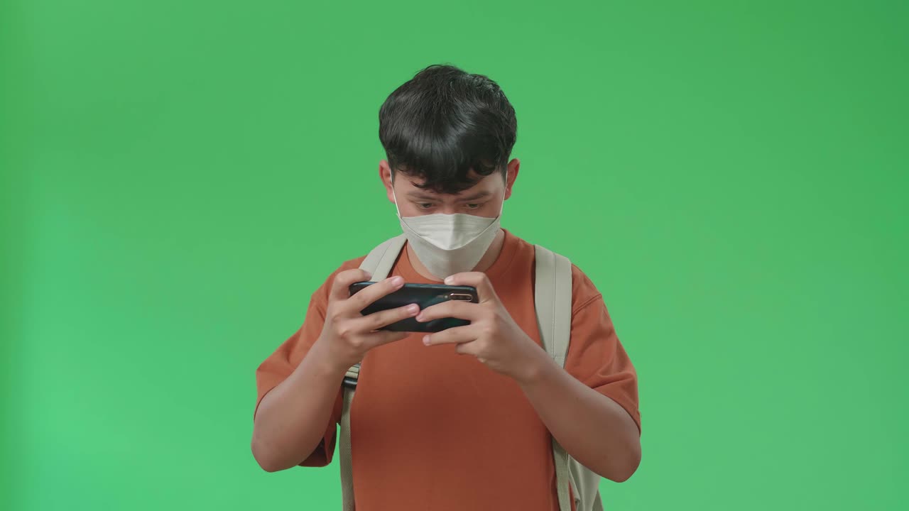 The Front View Of Asian Boy Student Wearing A Mask, Backpack And Brown T-Shirt Playing Game On Mobile Phone While Walking To School On Green Screen Chroma Key.