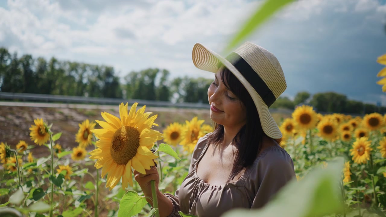 Primo piano di una donna con un cappello a tesa larga in piedi tra i girasoli, che tiene in mano ed esamina i fiori. La scena presenta vivaci fioriture gialle con foglie verdi sotto cieli azzurri e limpidi, creando un'atmosfera vibrante.