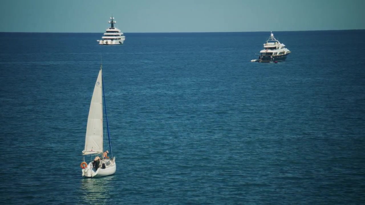 Barca a vela che naviga nel profondo blu del Mar Mediterraneo vicino ad Antibes, Costa Azzurra, con uno yacht di lusso visibile in lontananza in una luminosa giornata estiva, Antibes, Francia.