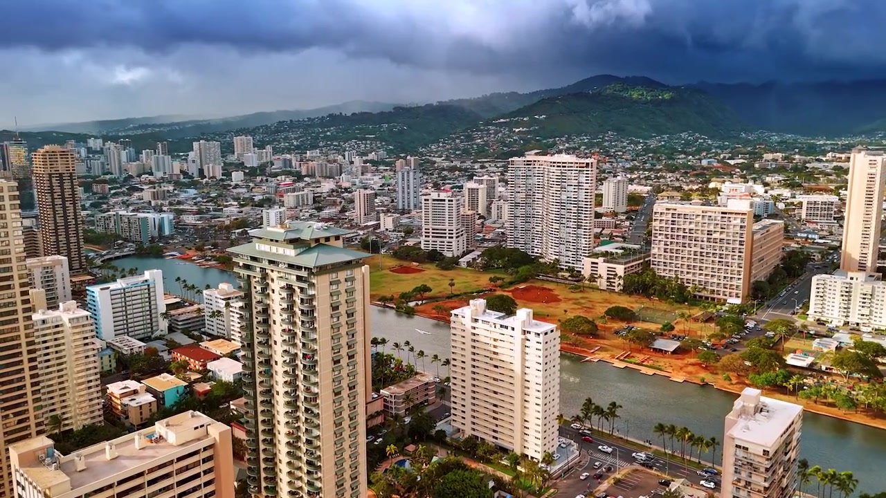 Volo tra le cime dei grattacieli di Waikiki in avvicinamento al canale Ala Wai. Vista panoramica di Honolulu con montagne coperte da nuvole scure sullo sfondo