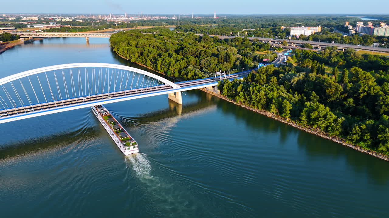 Lunga imbarcazione fluviale che viaggia lungo il Danubio a Bratislava, Slovacchia. Il veicolo acquatico passa sotto il Ponte Appolo. Vista aerea sulla lussureggiante vegetazione sul lungofiume.